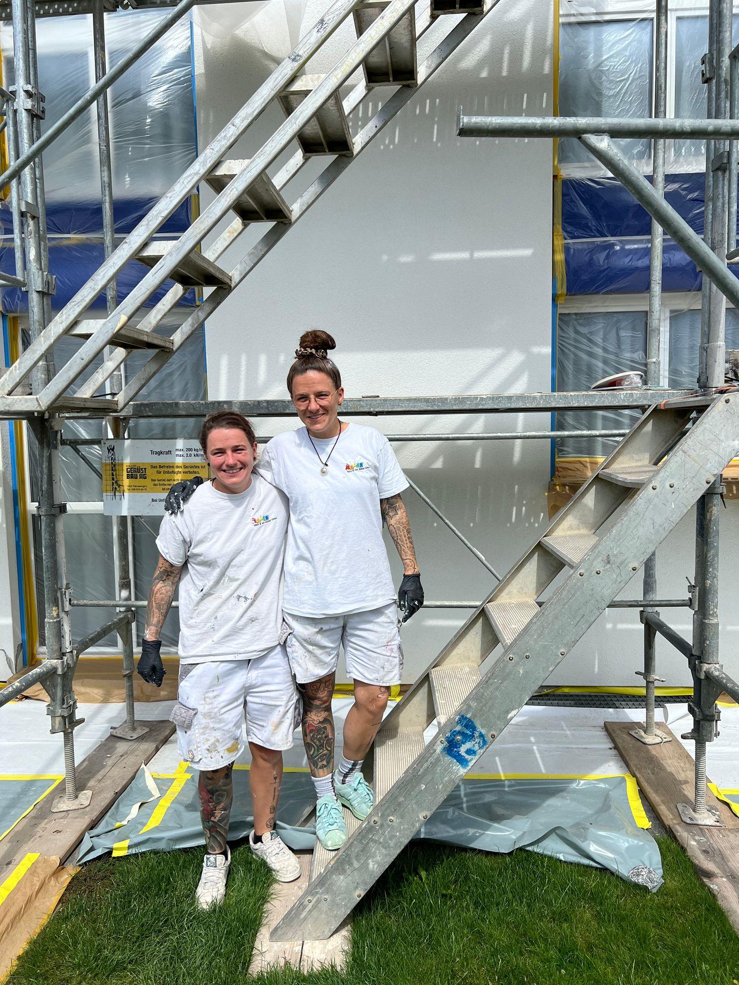 Two individuals in white painting uniforms standing together under a metal scaffold at a construction site.