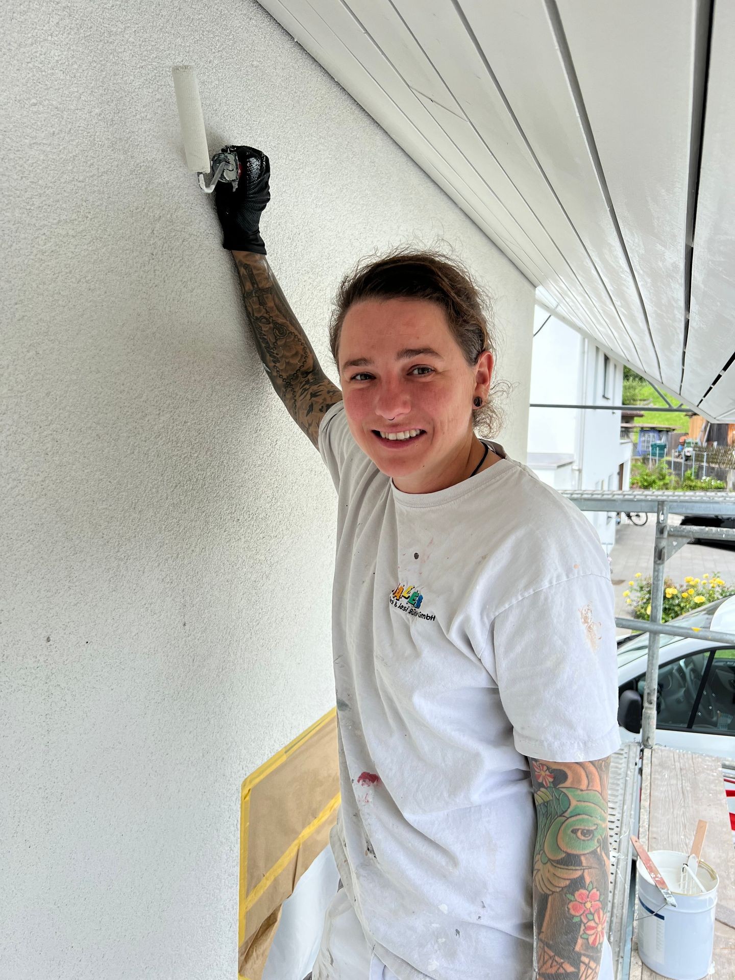 Person in white clothing with tattoos painting a wall with a roller brush, standing on scaffolding.