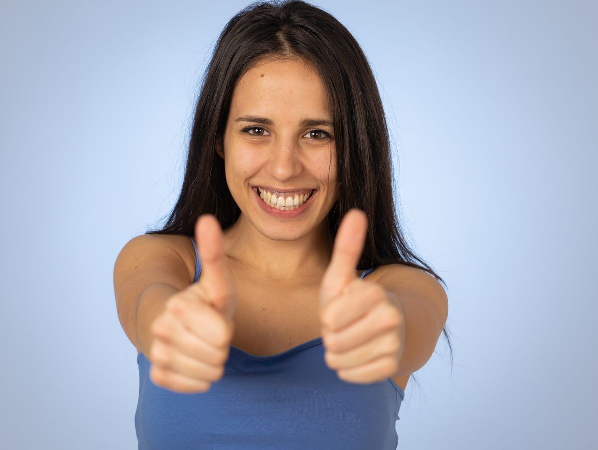 Portrait of beautiful young latin woman showing thumbs up sign feeling cheerful, happy and satisfied. Young happy student woman making thumb up in joyful approval gesture isolated on blue background.